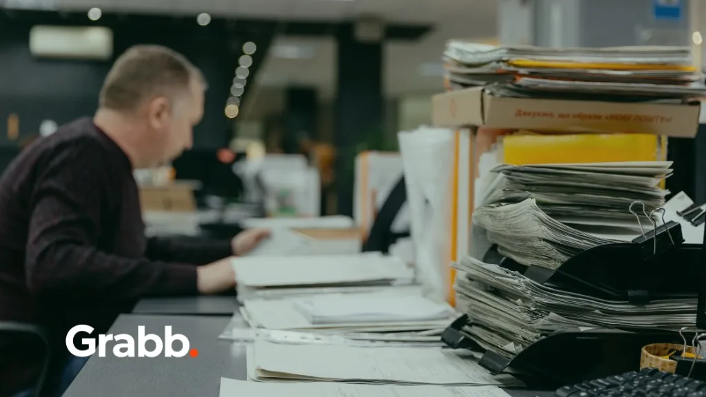 Stacks of clipped purchase orders and paperwork on a desk beside a keyboard, with an office worker reviewing documents in the background