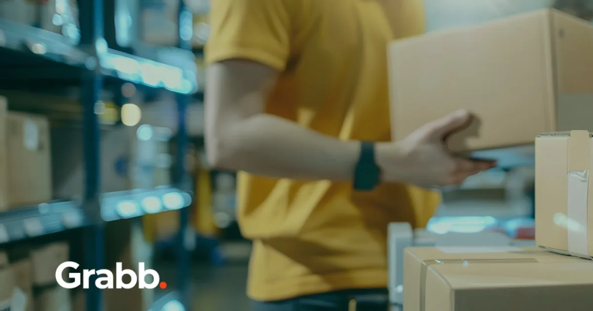 Warehouse associate in a yellow shirt holding a cardboard shipping box in front of blue storage racks